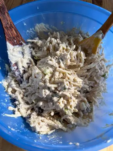 A large blue plastic bowl containing shredded hash browns, cheese, green onions and a liquid mixture containing cream of mushroom soup. There are 2 cooking utensils resting in the bowl.