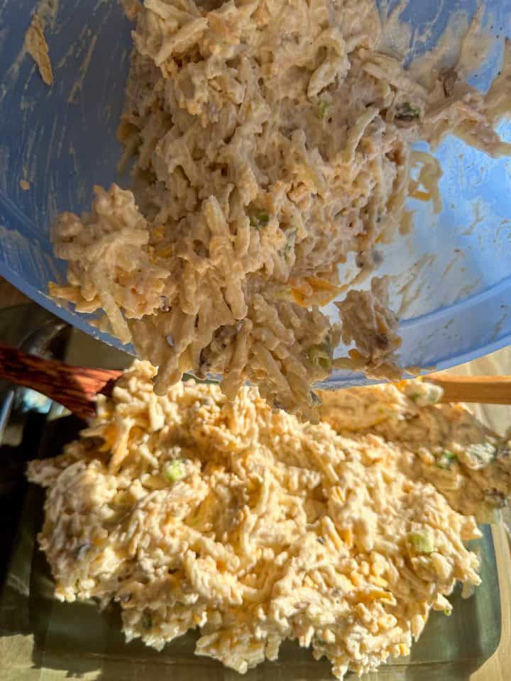 A large blue bowl containing shredded hash browns, cheese, green onions and a liquid mixture containing cream of mushroom soup poised above a casserole dish. The mixture is being transferred from the bowl to the casserole dish.