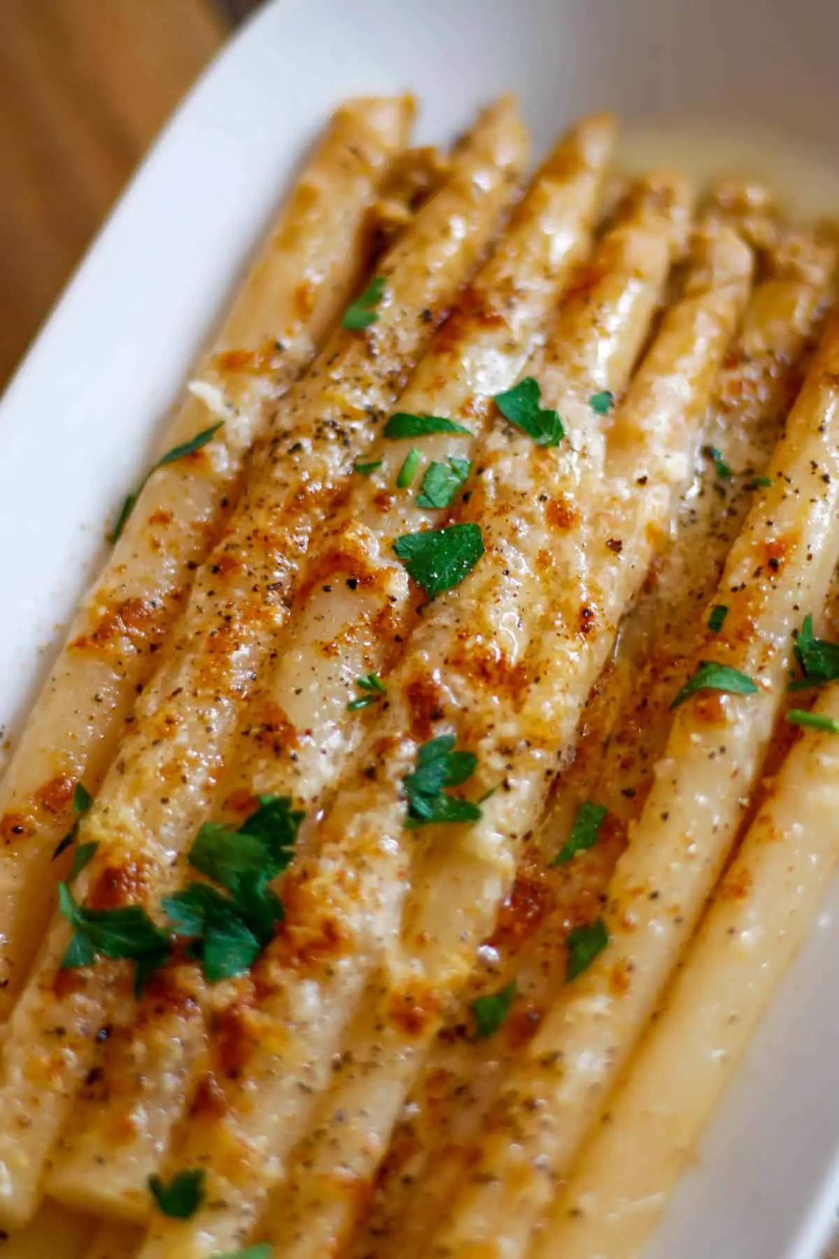 White asparagus with melted and browned cheese garnished with Italian parsley in a white dish.