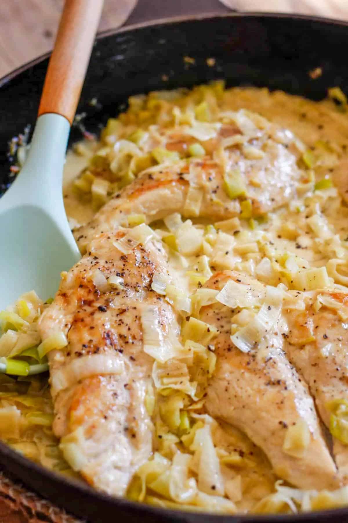 A cast iron pan containing creamy chicken breasts and leeks. There is a blue spoon with a wooden handle in the bowl.