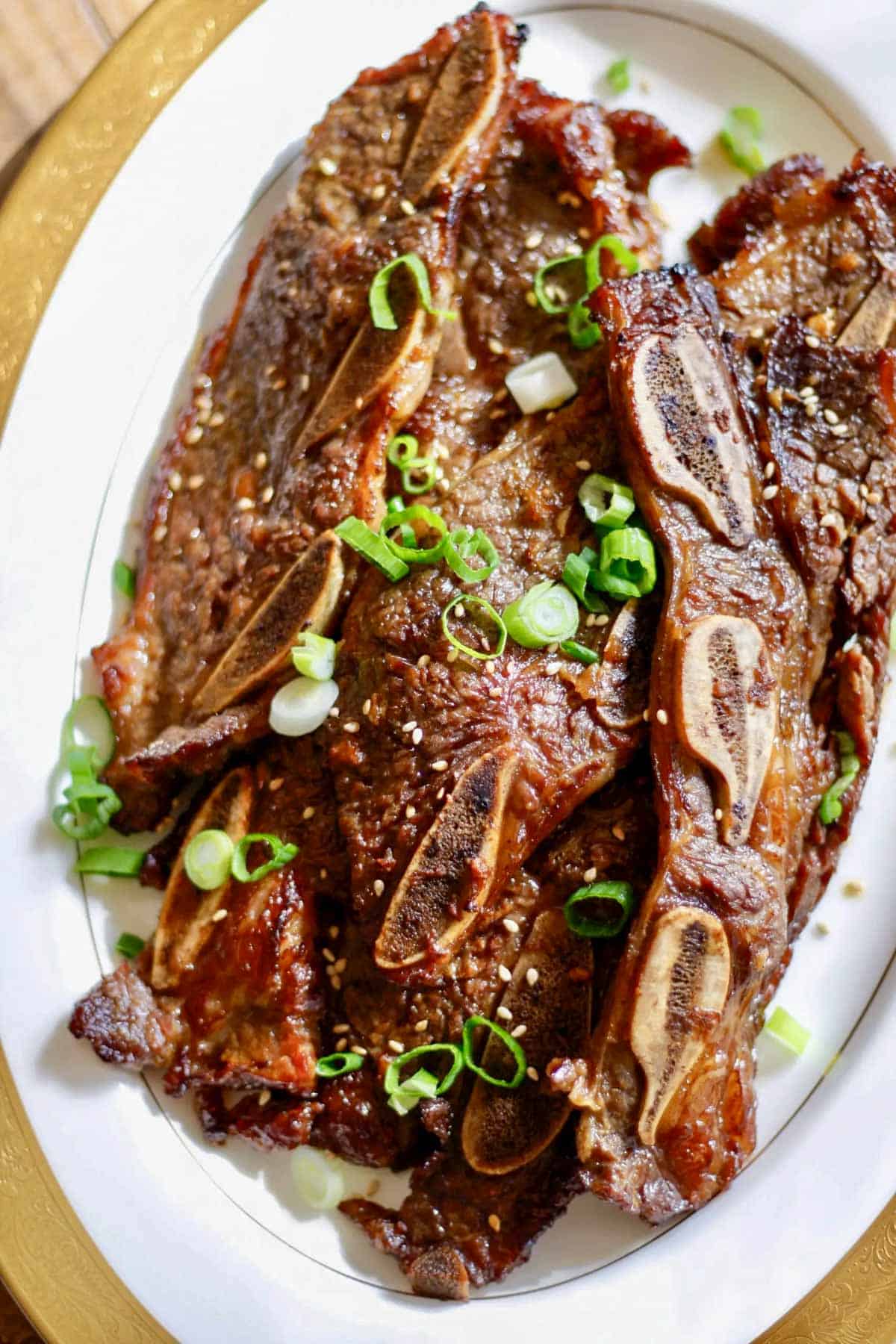 A gold rimmed serving dish containing Korean Short Ribs garnished with sesame seeds and green onions.