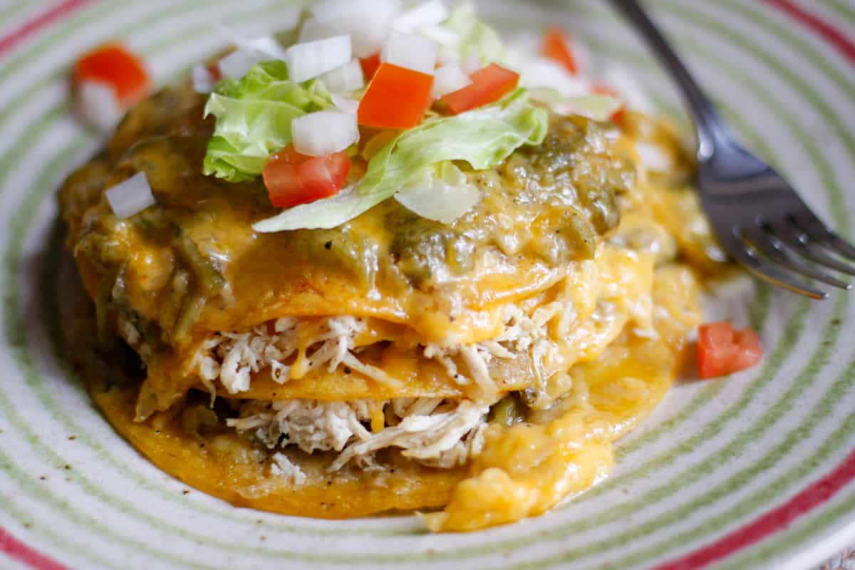 Corn tortillas dipped in green chile sauce stacked on top of each other and filled with shredded chicken and cheese. The cheese is melted and the stack of enchiladas is topped with melted cheese and green chile sauce, garnished with chopped tomatoes and onions, and shredded lettuce. There is a fork on the side of the plate.