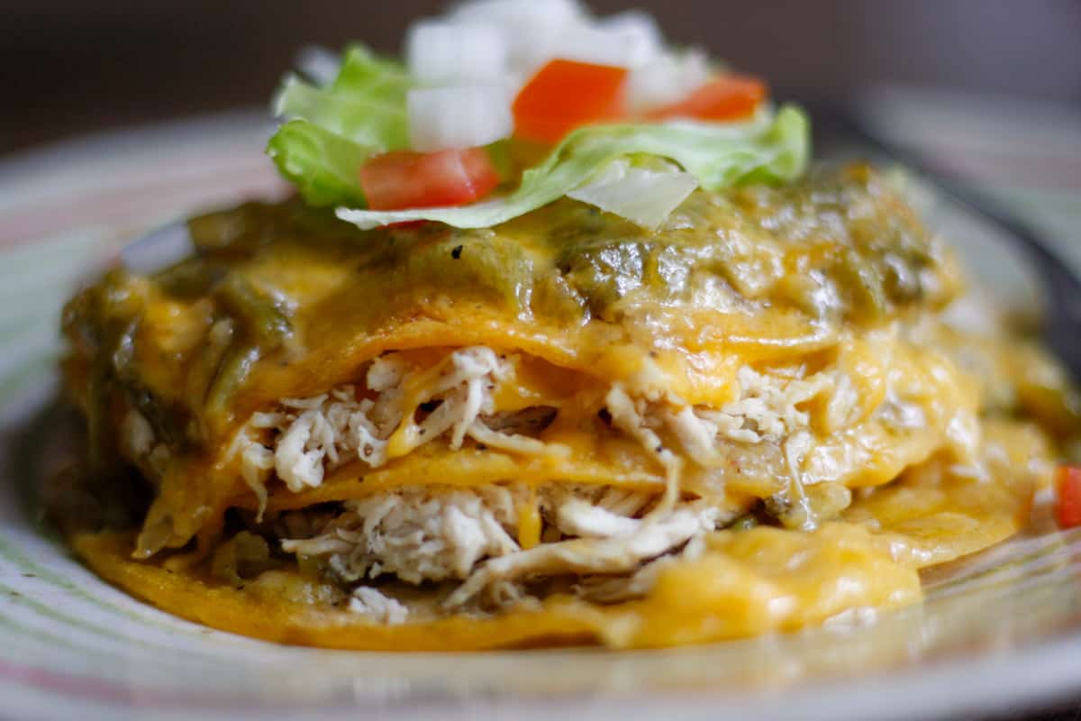Corn tortillas dipped in green chile sauce stacked on top of each other and filled with shredded chicken and cheese. The cheese is melted and the stack of enchiladas is topped with melted cheese and green chile sauce, garnished with chopped tomatoes and onions, and shredded lettuce.