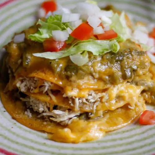 Corn tortillas dipped in green chile sauce stacked on top of each other and filled with shredded chicken and cheese. The cheese is melted and the stack of enchiladas is topped with melted cheese and green chile sauce, garnished with chopped tomatoes and onions, and shredded lettuce.