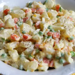 Belizean Potato Salad With Peas And Carrots with black pepper sprinkled on top in a white bowl.