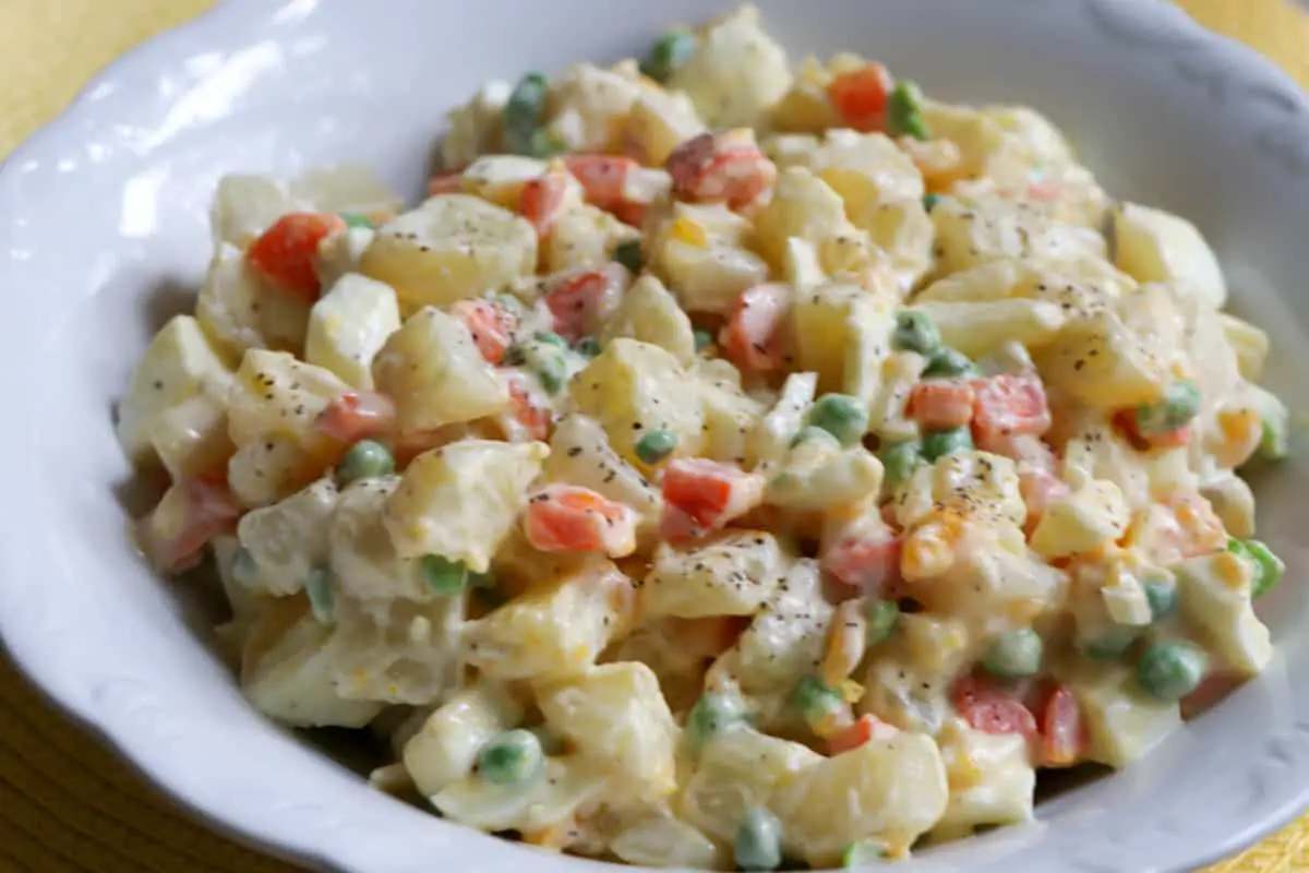 Belizean Potato Salad With Peas And Carrots with black pepper sprinkled on top in a white bowl.