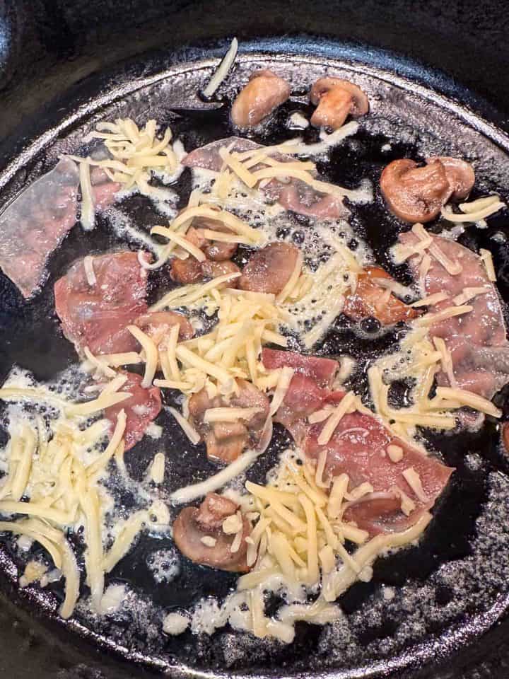 A cast iron skillet with melted butter, slices of prosciutto, mushrooms, and shredded gouda.