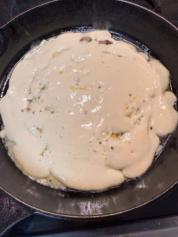 A cast iron pan containing a pancake that is cooking. The top is wet with bubbles forming and is just about ready to turn.