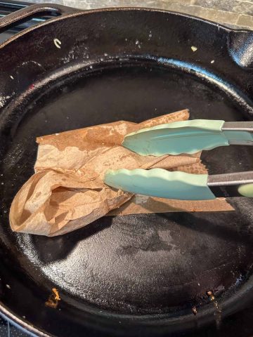 A cast iron skillet being wiped and cleaned with a paper towel being moved by a pair of tongs.
