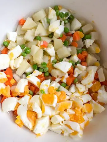 A white bowl containing chopped hard boiled egg, cut up pieces of boiled potato, diced carrots, and green peas.