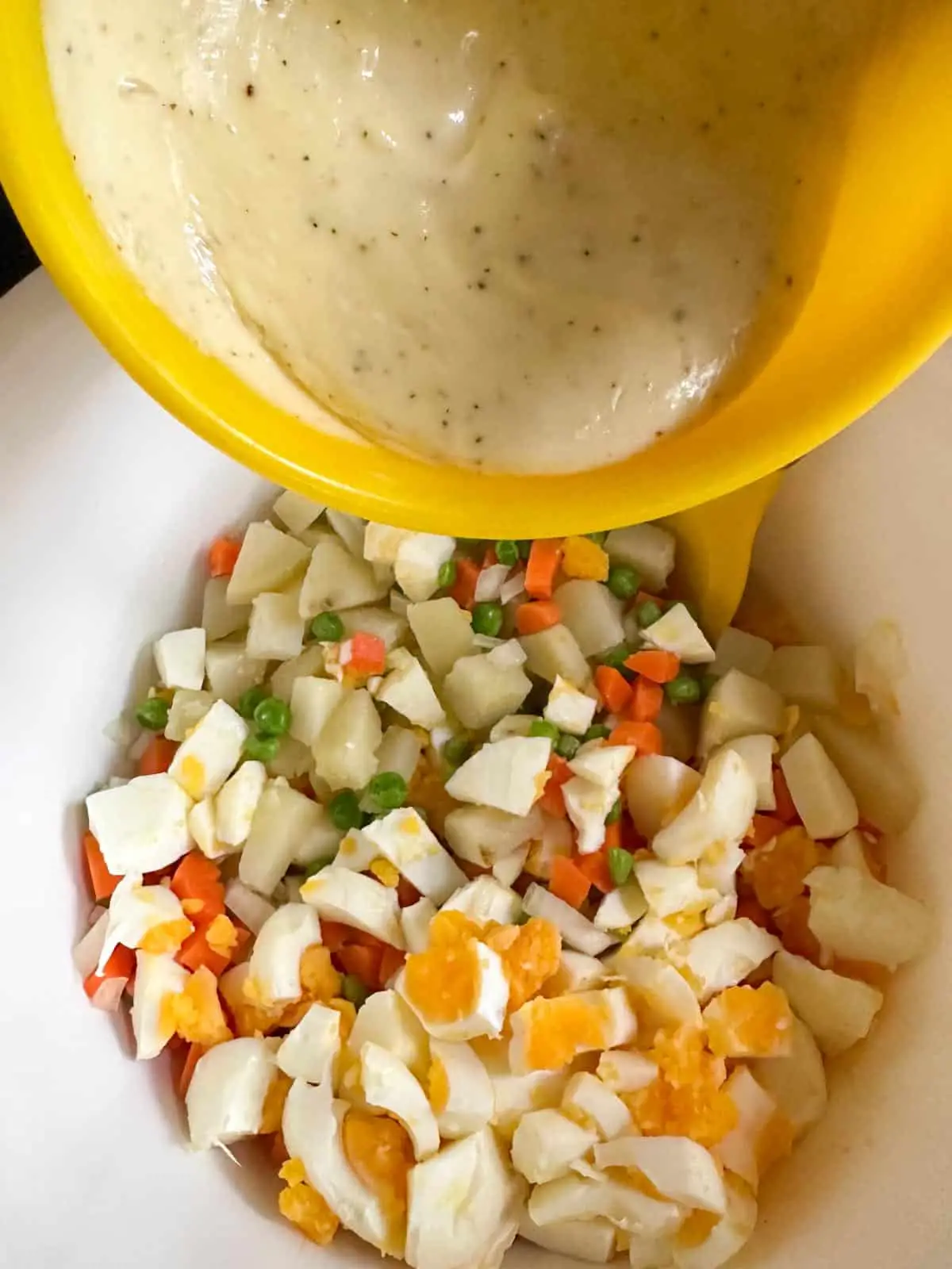 A white bowl containing chopped hard boiled egg, cut up pieces of boiled potato, diced carrots, and green peas. There is a yellow bowl containing a salad cream and mayo mixture poised over the white bowl.