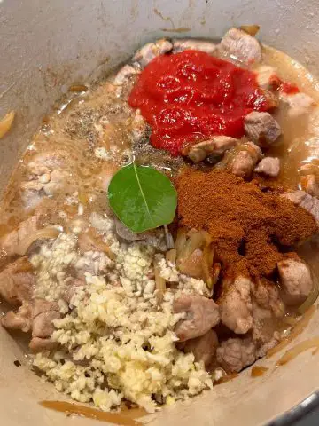 Tomatoes, bay leaf, paprika, seasonings, and minced garlic added to chunks of pork being cooked in broth in a Dutch oven.
