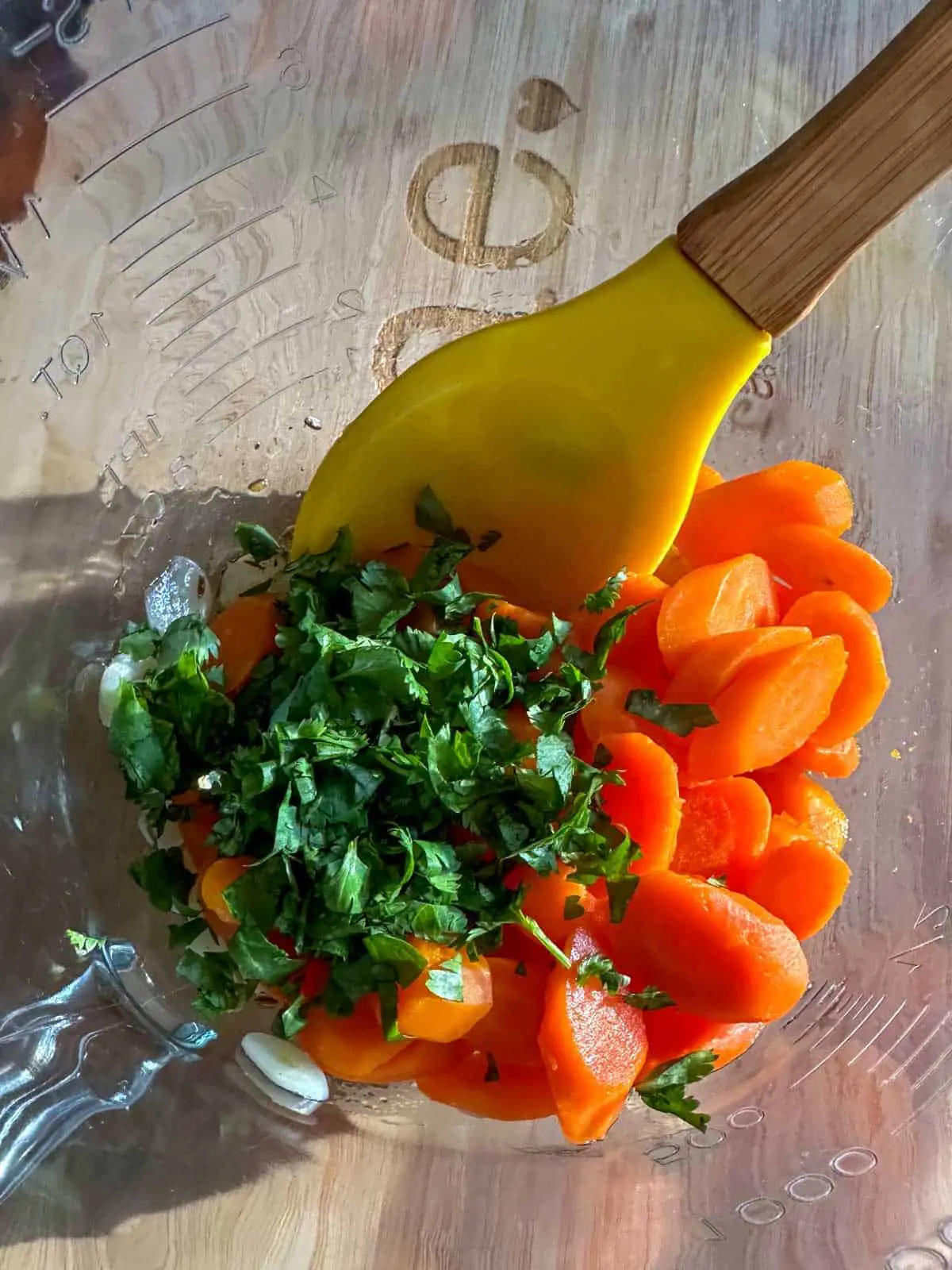 Sliced carrots, sliced garlic, and finely chopped cilantro in a glass bowl with a yellow spoon.