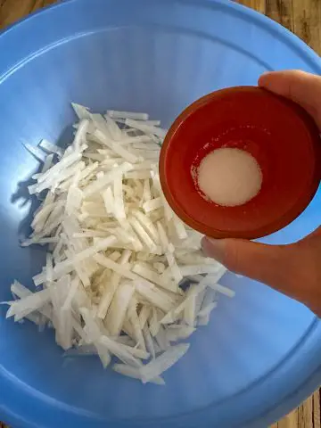 A blue bowl containing julienned daikon radish. A hand is holding a small bowl with salt above the blue bowl.