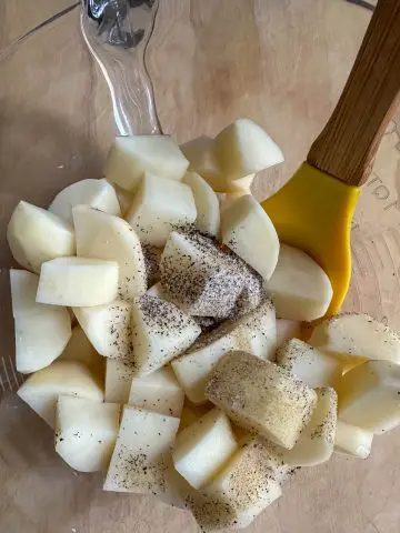 Cut up pieces of russet potato sprinkled with salt and pepper in a glass bowl with a yellow spoon with wooden handle resting in the bowl.