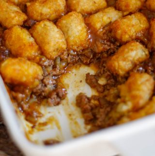 A white casserole dish containing a tater tot casserole. Part of the casserole has been removed so that the inside part of the casserole can be seen.