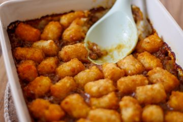A casserole dish containing a tater tot casserole with ground beef. There is a blue spoon resting in the dish.
