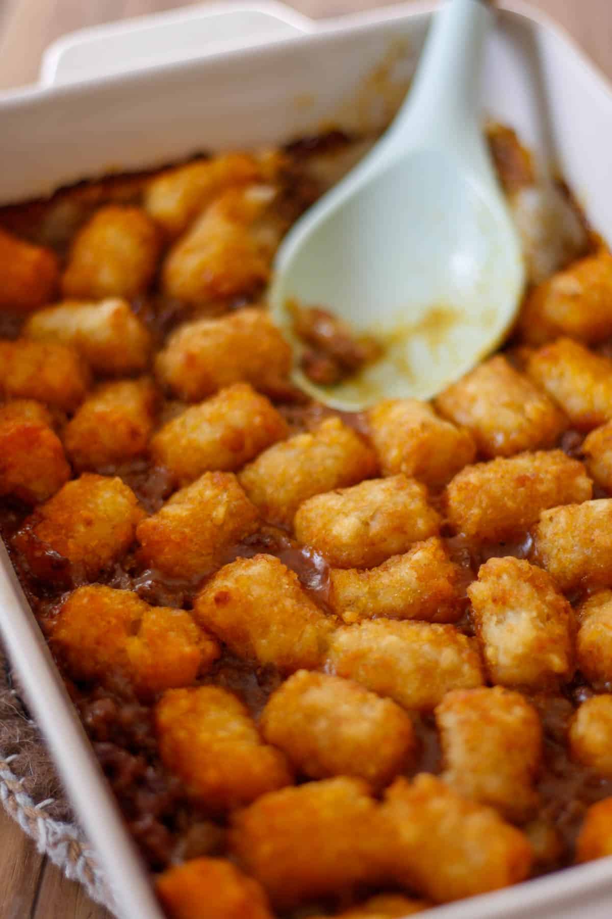 A casserole dish containing a tater tot casserole with ground beef. There is a blue spoon resting in the dish.