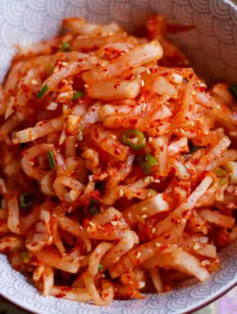 Korean radish salad which is julienned daikon radish in a spicy red sauce garnished with green onions and sesame seeds in a bowl.