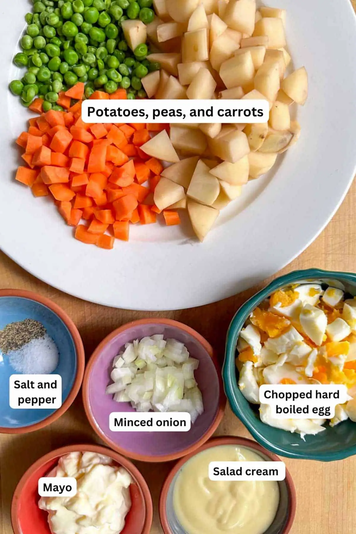 A white plate with pieces of potato and carrot cut into small pieces, and sweet peas; bowls with salt and pepper, minced onion, chopped hard boiled egg, mayo and salad cream.