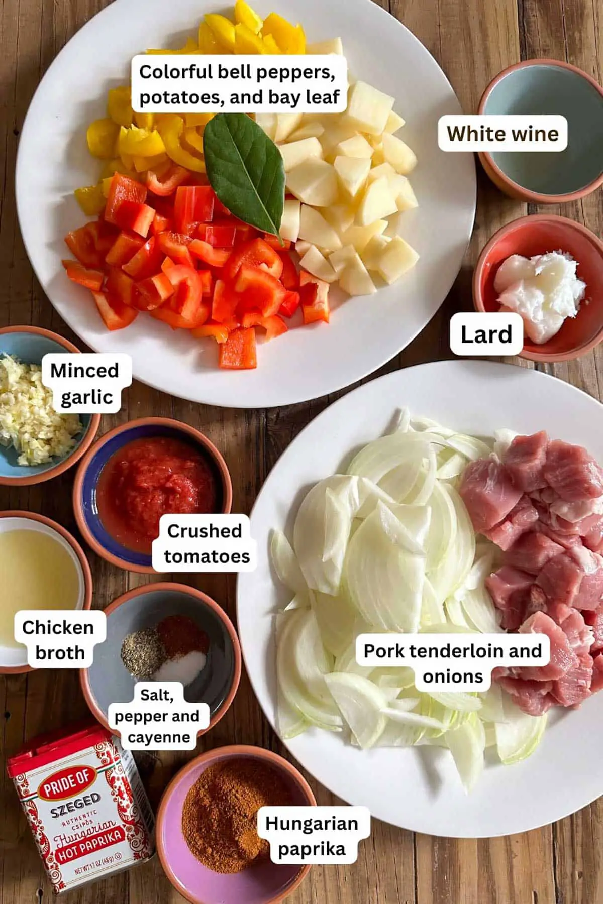 Ingredients for Hungarian pork goulash displayed on plates and bowls including: bell peppers, potatoes, bay leaf, white wine, lard, minced garlic, crushed tomatoes, chicken broth, seasonings, paprika, pork tenderloin, sliced onions, and a container of Hungarian paprika.