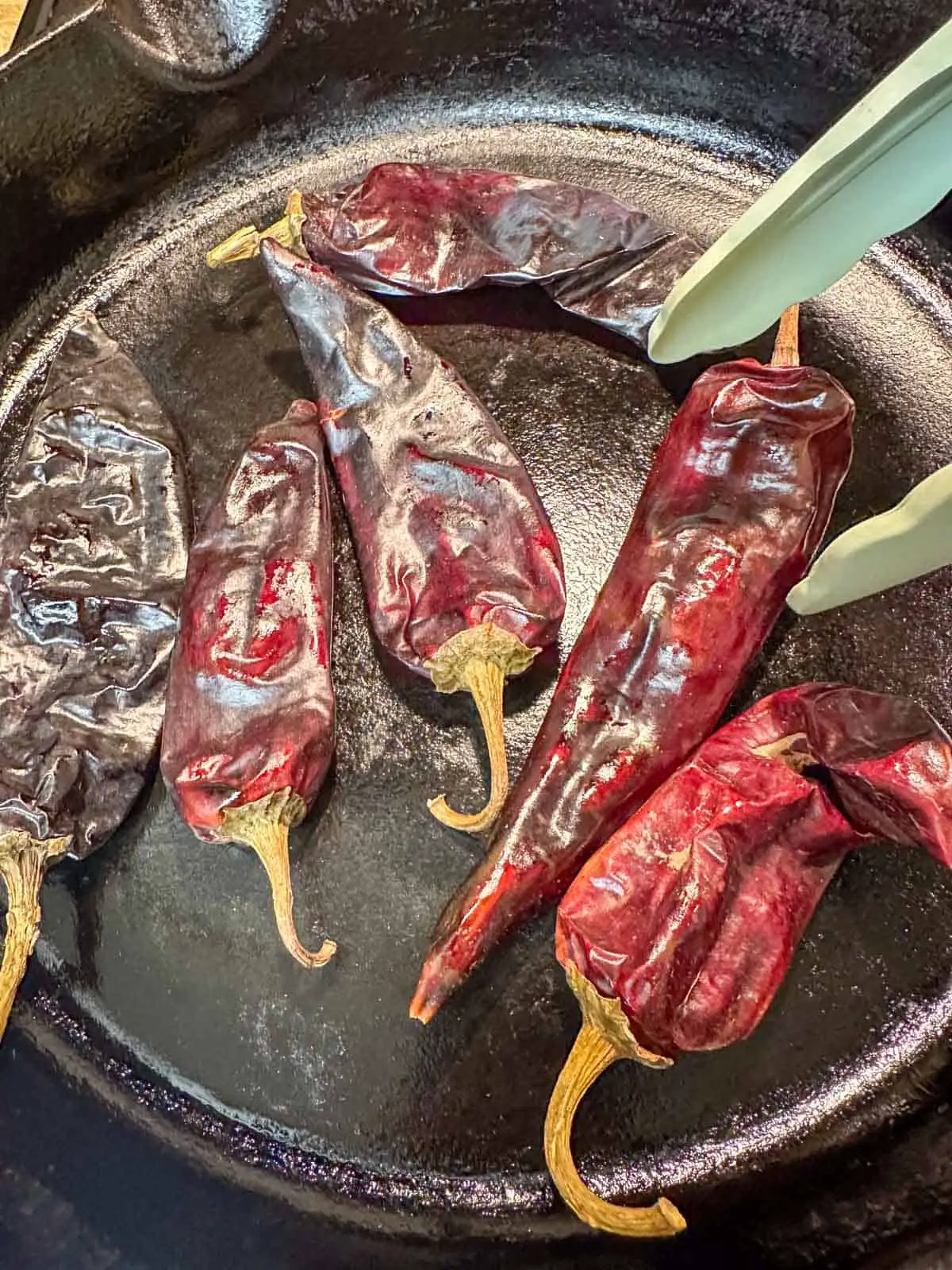 New Mexico red chile pods toasting in a cast iron pan. There is a set of tongs poised over one of the chile pods.