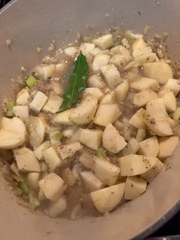 A Dutch oven with cut up potatoes, celery, and minced onion in broth. There is a bay leaf on top of these ingredients.