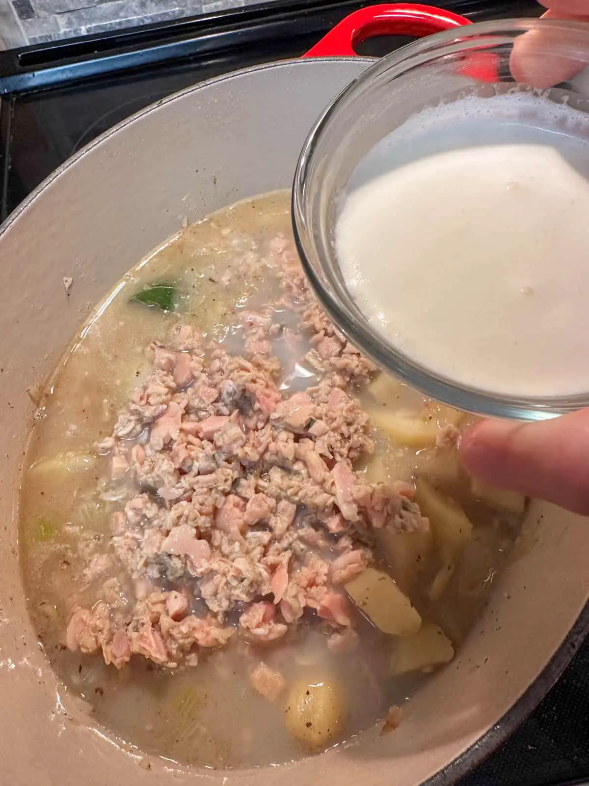Minced clams and potatoes in broth in a Dutch oven. There is a person holding a small bowl with cream above the Dutch oven.