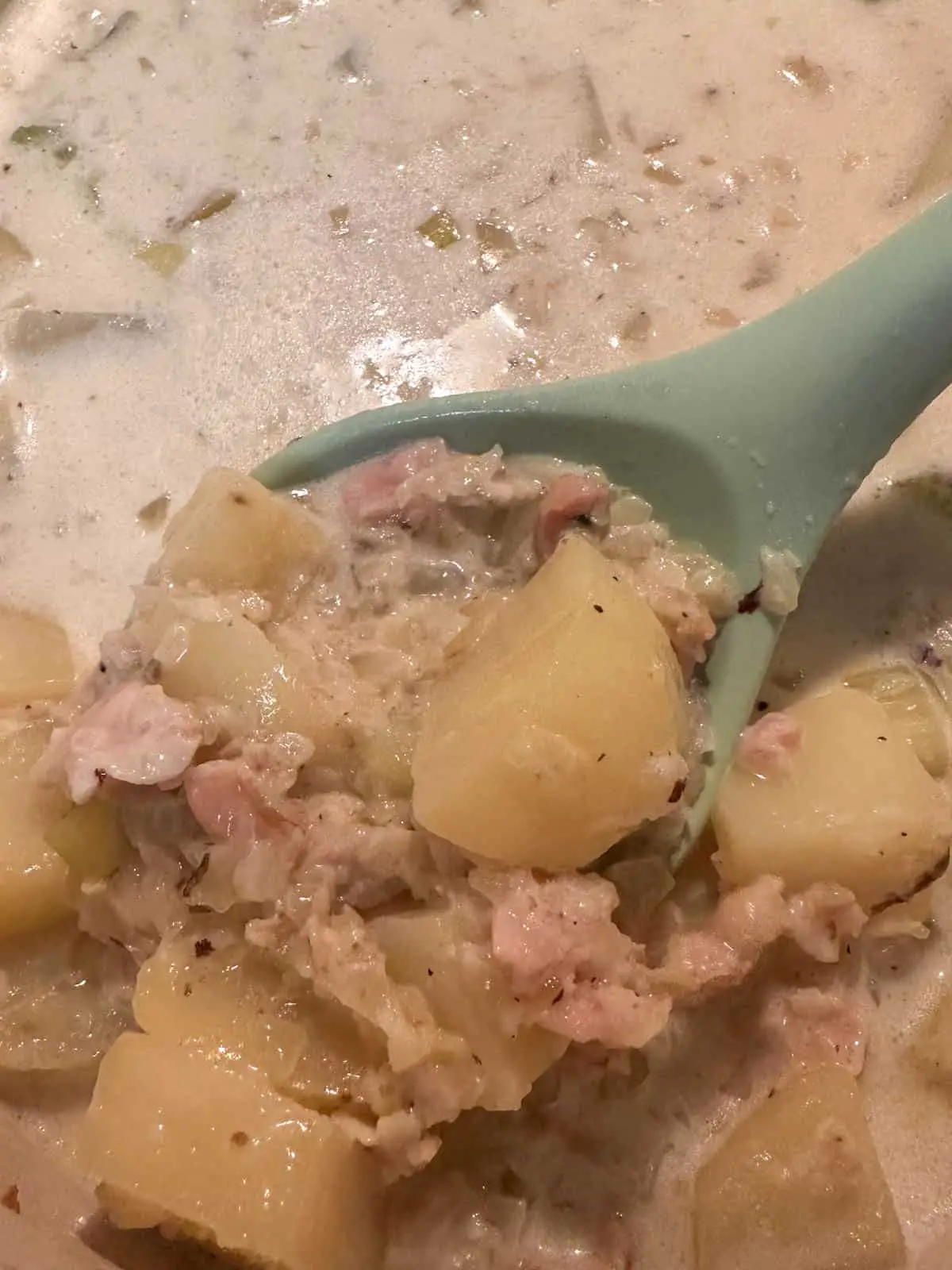New England Clam Chowder in a Dutch oven. There is a blue spoon containing some of the chowder showing off clams and potatoes.