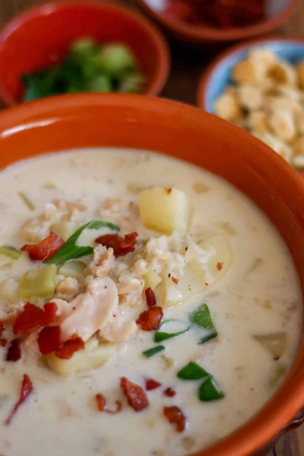 New England Clam Chowder in a terracotta bowl garnished with bacon and green onions. There are bowls containing green onions, oyster crackers, and bacon bits in the background.