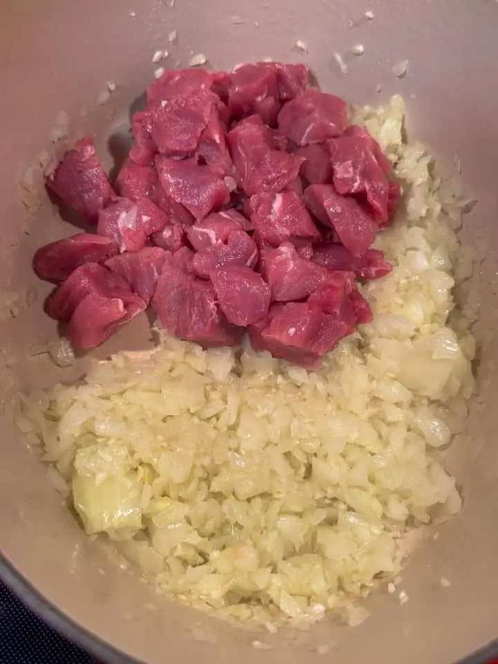 Pieces of uncooked pork tenderloin and diced onion and garlic in a Dutch oven.
