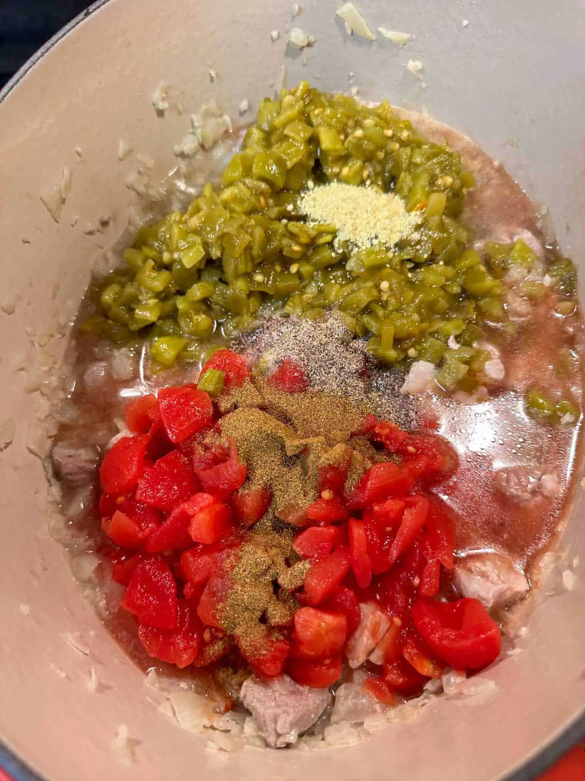 Chicken broth, green chiles, diced Chicken broth, green chiles, diced tomatoes and onion, and pork tenderloin in a Dutch oven. Seasonings including chicken bouillon, salt and pepper, and green chile powder have been added to the top of the other ingredients.