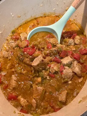 Colorado Green Chili which is pork tenderloin pieces, diced tomatoes and green chilis in a Dutch oven, with a blue spoon with wooden handle resting in the Dutch oven.