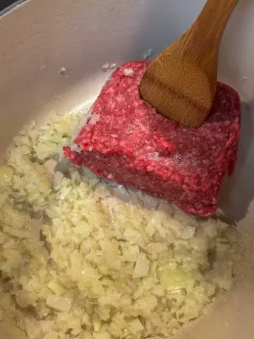 Finely chopped onion, garlic and shallot and a block of beef mince in a Dutch oven. There is a wooden spoon inserted in the block of beef mince.