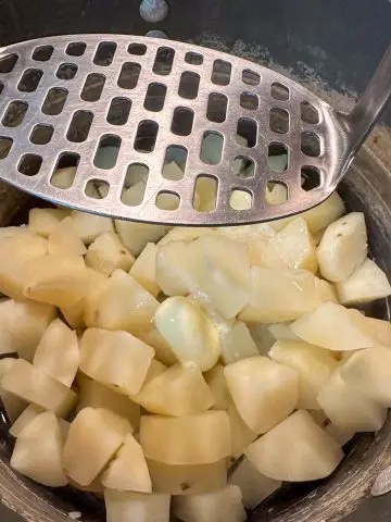 Cut up pieces of cooked Russet potatoes with a potato masher poised above the potatoes.