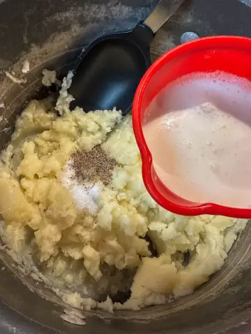 Mashed potatoes with salt and pepper in a saucepan. There is a black spoon in the saucepan and a red cup with heavy cream poised over the saucepan.