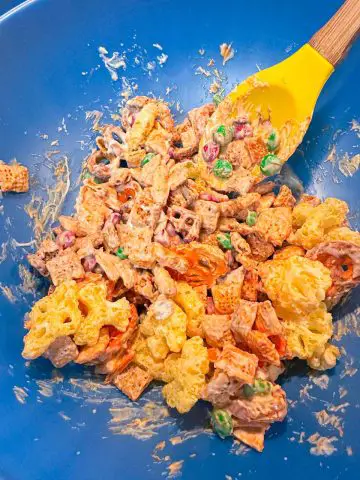 Red and green M&M's, Chex Mix, Cheetos Pretzels and Cheetos snowflakes tossed with melted white chocolate in a large blue bowl. There is a yellow spoon with a wooden handle in the bowl.