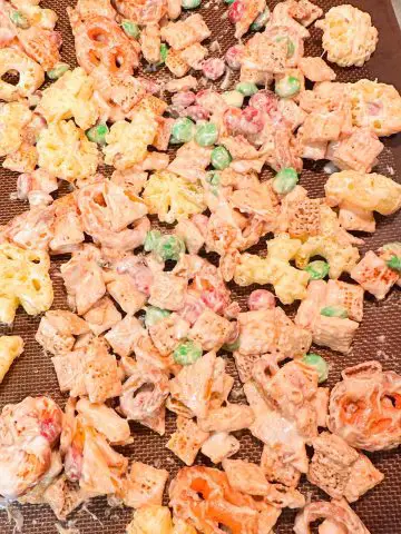 Red and green M&M's, Chex Mix, Cheetos Pretzels and Cheetos snowflakes tossed with melted white chocolate spread out on a silicone mat.
