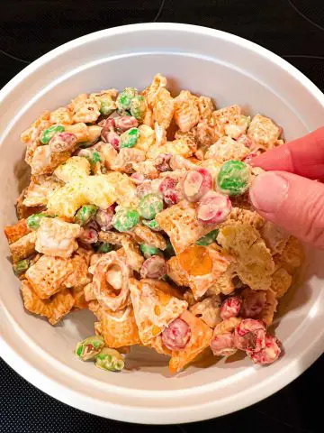 A white bowl containing Christmas White Chocolate Cheddar Texas Trash. There is a glimpse of someone holding a piece of the Texas Trash containing white chocolate covered M&M's in the picture.