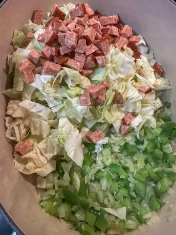 A Dutch oven containing celery, green bell pepper, onion, chopped cabbage and Andouille sausage.