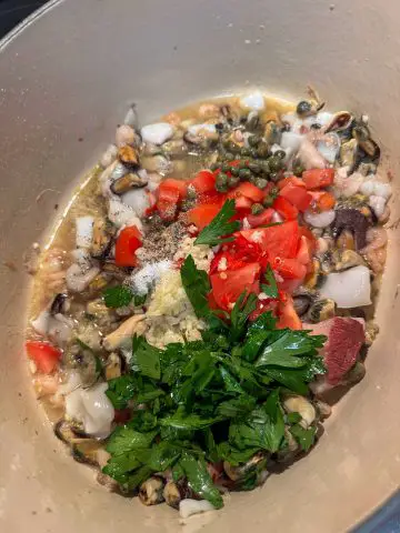 A Dutch oven containing seafood mix, Italian parsley, chopped tomato, salt and pepper, garlic, capers, and broth.