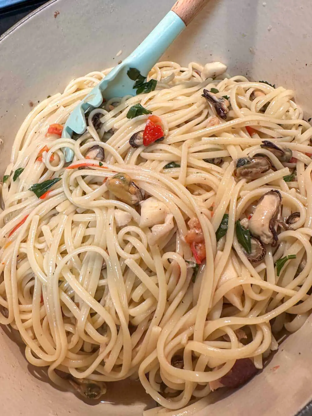 Cooked linguine with seafood mix, chopped tomato and Italian parsley in a Dutch oven. There is a blue pasta spoon with wooden handle resting in the pasta.