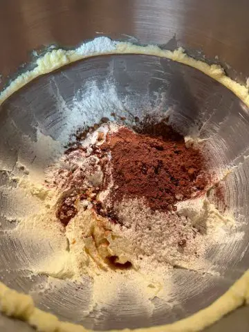 A stainless steel mixing bowl with creamed butter and sugar. Cocoa powder, flour and vanilla have been added to the creamed butter and sugar.