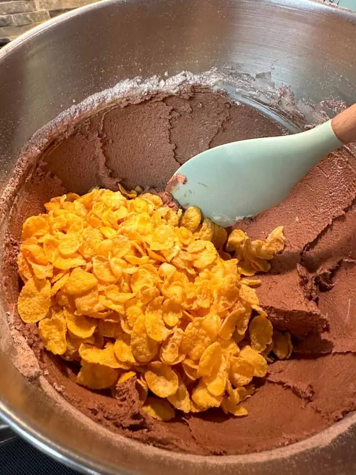 A stainless steel mixing bowl with creamy chocolate cookie dough and corn flakes. There is a blue spatula with a wooden handle resting in the bowl.