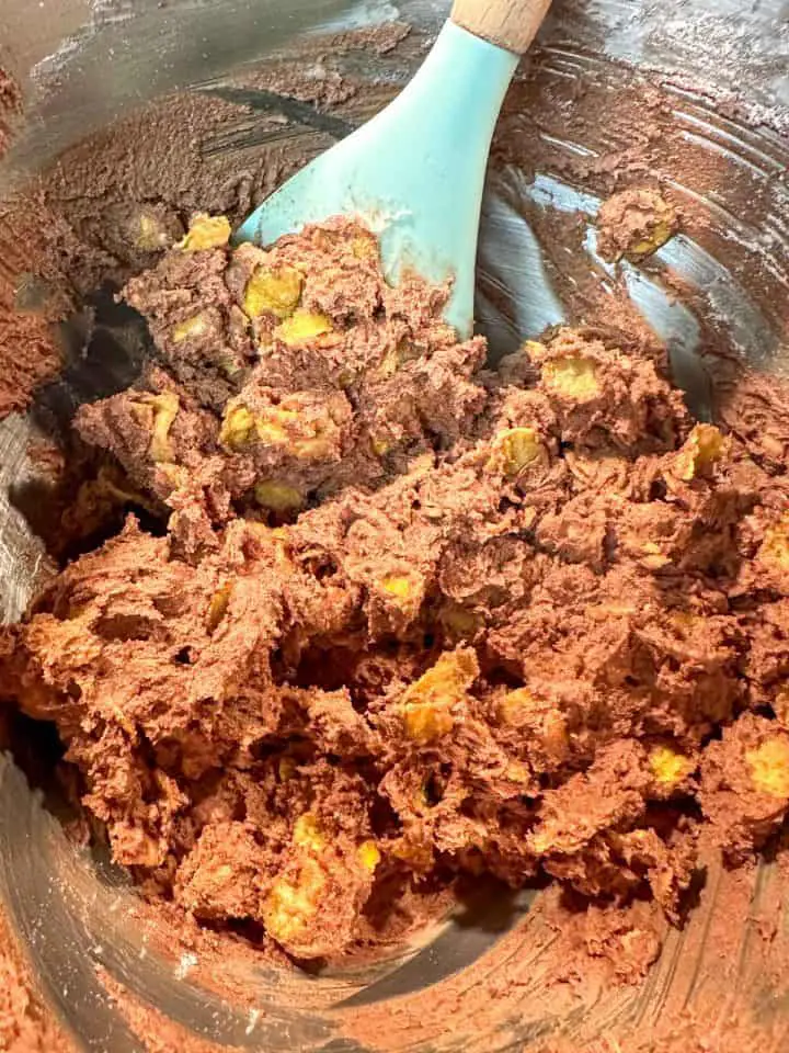 A stainless steel mixing bowl with creamy chocolate cookie dough and corn flakes mixed into the dough. There is a blue spatula with wooden handle resting in the bowl.