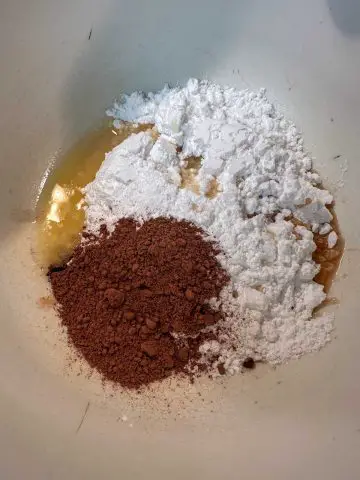 A mixing bowl containing powdered sugar, melted butter, vanilla, and cocoa powder.