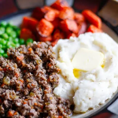 Ground beef also called mince, mashed potatoes also called tatties with melted butter, roasted carrots, and peas on a plate.