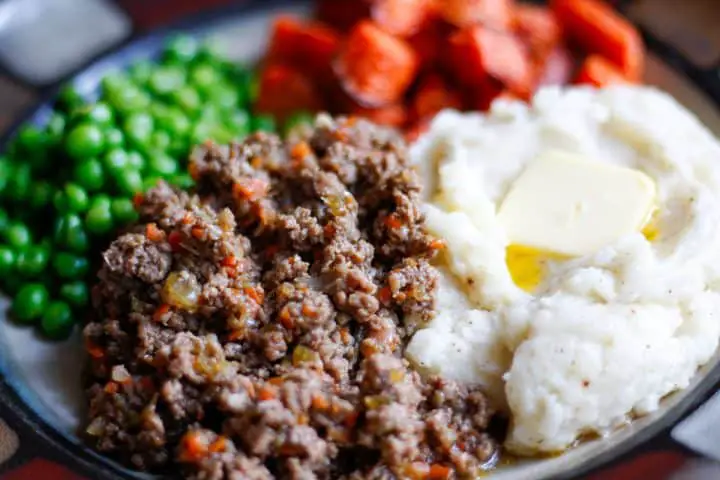 Ground beef also called mince, mashed potatoes also called tatties with melted butter, roasted carrots, and peas on a plate.