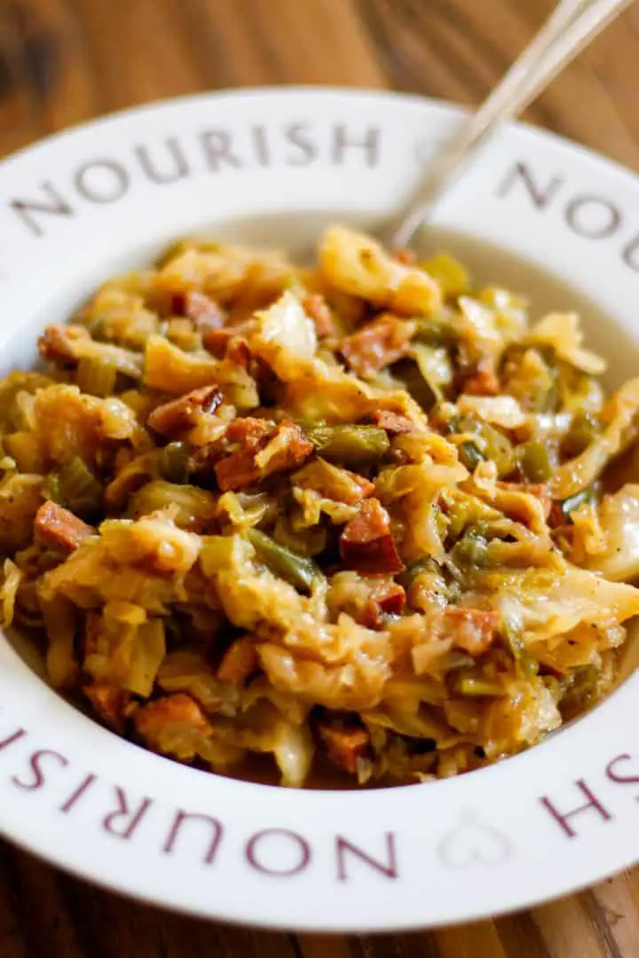 A bowl with the word "Nourish" on the rim containing Cajun smothered cabbage. There is a fork protruding from the bowl.