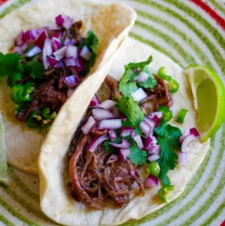 Barbacoa tacos garnished with cilantro, red onion, chilis, and lime on a plate with green and red concentric circles.